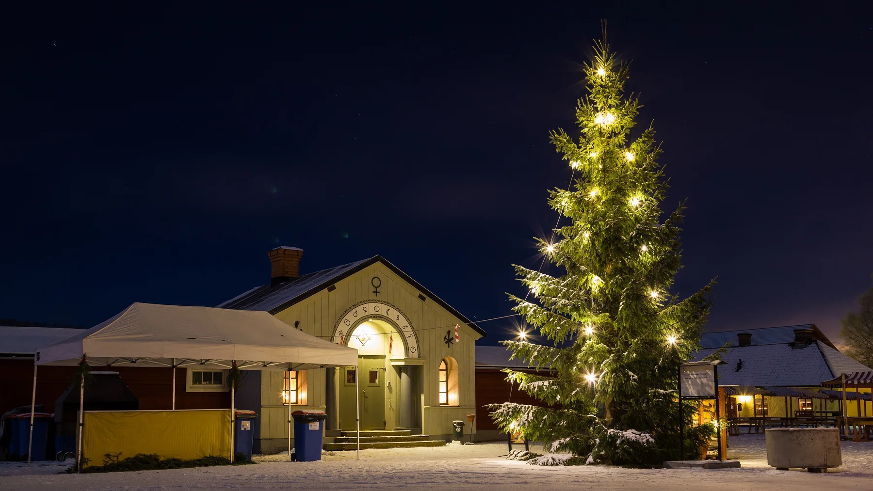 Upplyst gruvbyggnad och julgran i snö vid Falu gruva under mörk kvällshimmel.