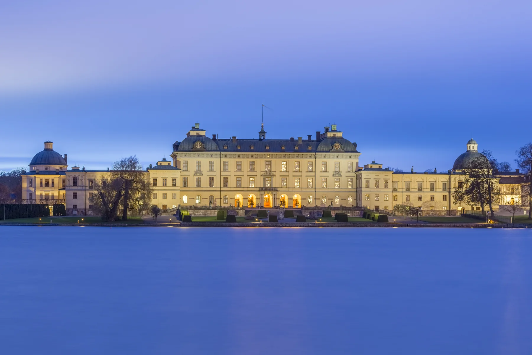 Drottningholms slott upplyst i skymning, sett över ett blått vinterlandskap vid vattnet.