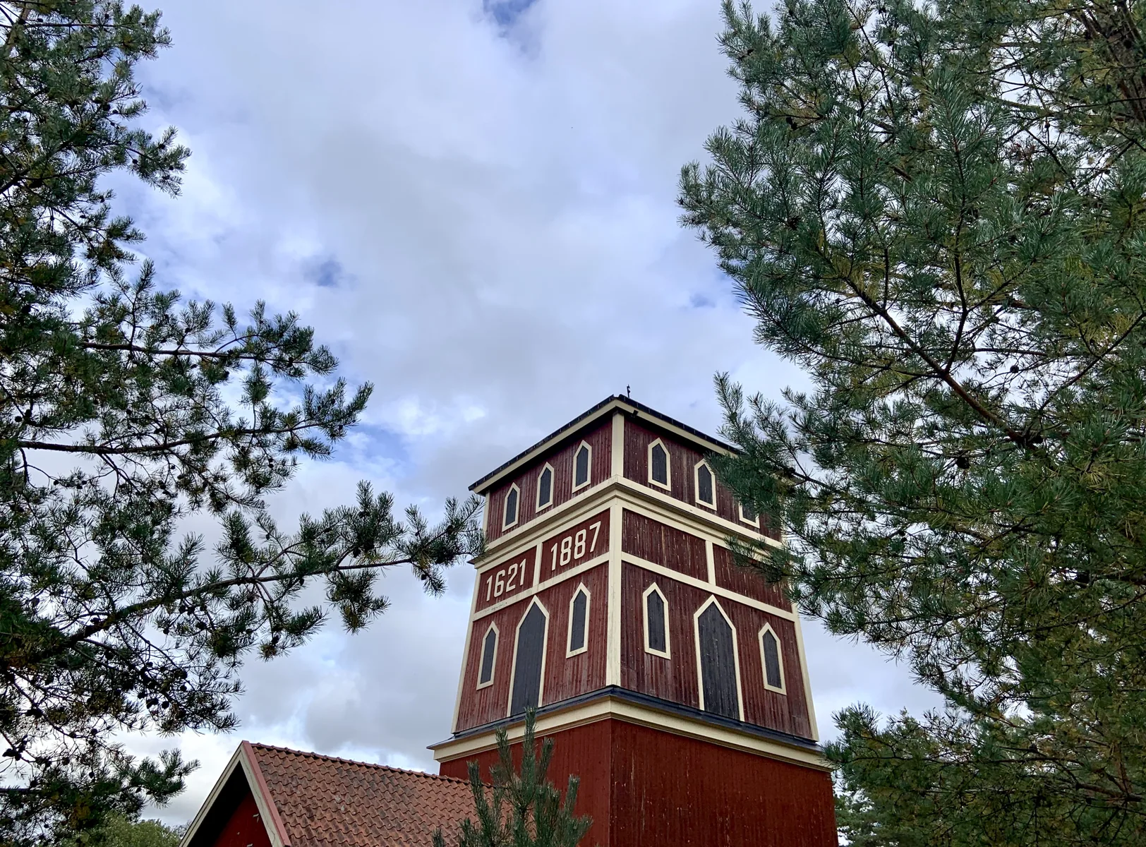 Rött schaktorn med årtalen 1621 och 1887, inramat av tallar och molnig himmel.